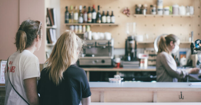 Two women wait for their order, being prepared by a sever wearing a face mask. Photo by Dan Burton on Unsplash.