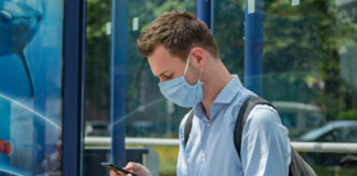 man with mask waits at bus stop.