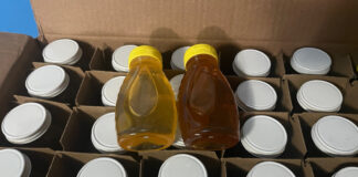 Two 8-ounce bottles of freshly harvested honey sitting on top of a box of two-dozen one-pound jars.